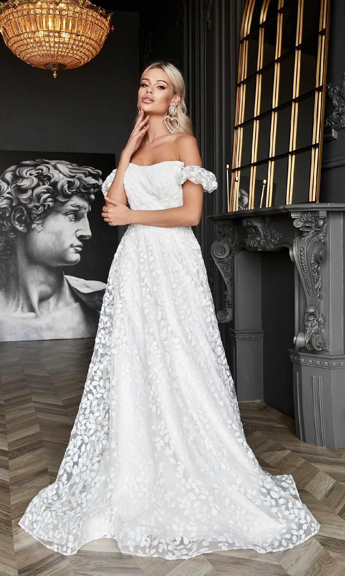 A woman wears the Long White Lace Formal Dress: Tina Holly TK023W, featuring floral patterns and an off-the-shoulder design, standing indoors near a fireplace with a statue and chandelier in the background.